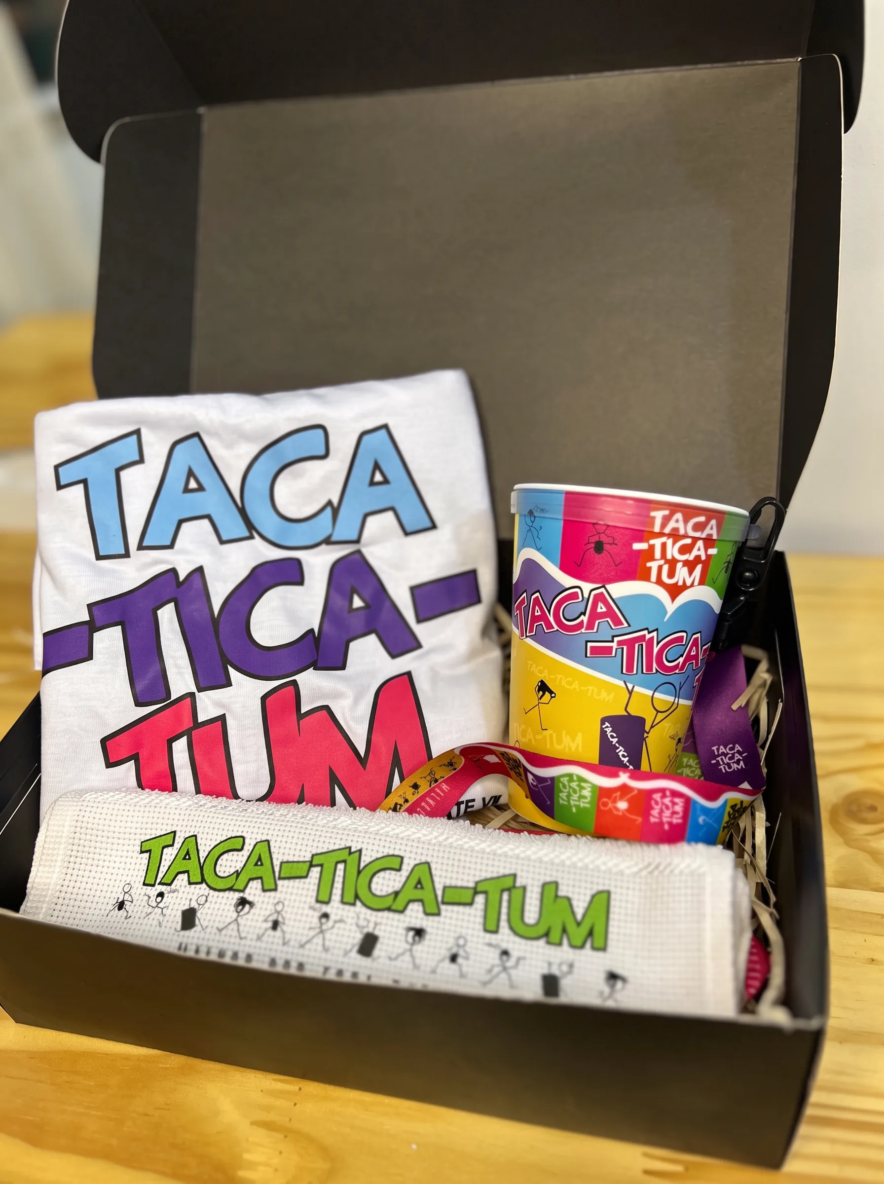 Caixa aberta com camiseta, copo, cordão e tecido TACATICATUM
